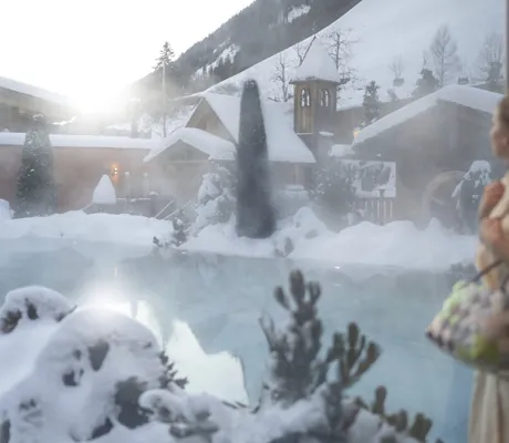 Frau im Bademantel mit Tasche blickt auf den dampfenden Außenpool des Hotel Quelle Nature Spa Resort in verschneiter Winterlandschaft mit Gebäuden und Bergen im Hintergrund