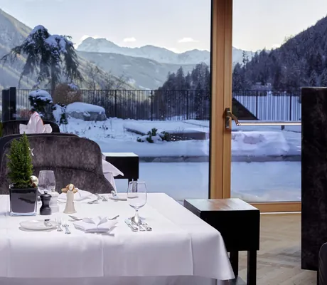 Eingedeckter Tisch im Restaurant des Hotel Quelle Nature Spa Resort mit weißer Tischdecke und Kerzenhalter vor großem Fenster mit Blick auf verschneite Berglandschaft