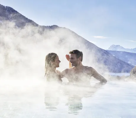 Paar im dampfenden Infinitypool des Hotel Quelle Nature Spa Resort mit Winter-Berglandschaft im Hintergrund