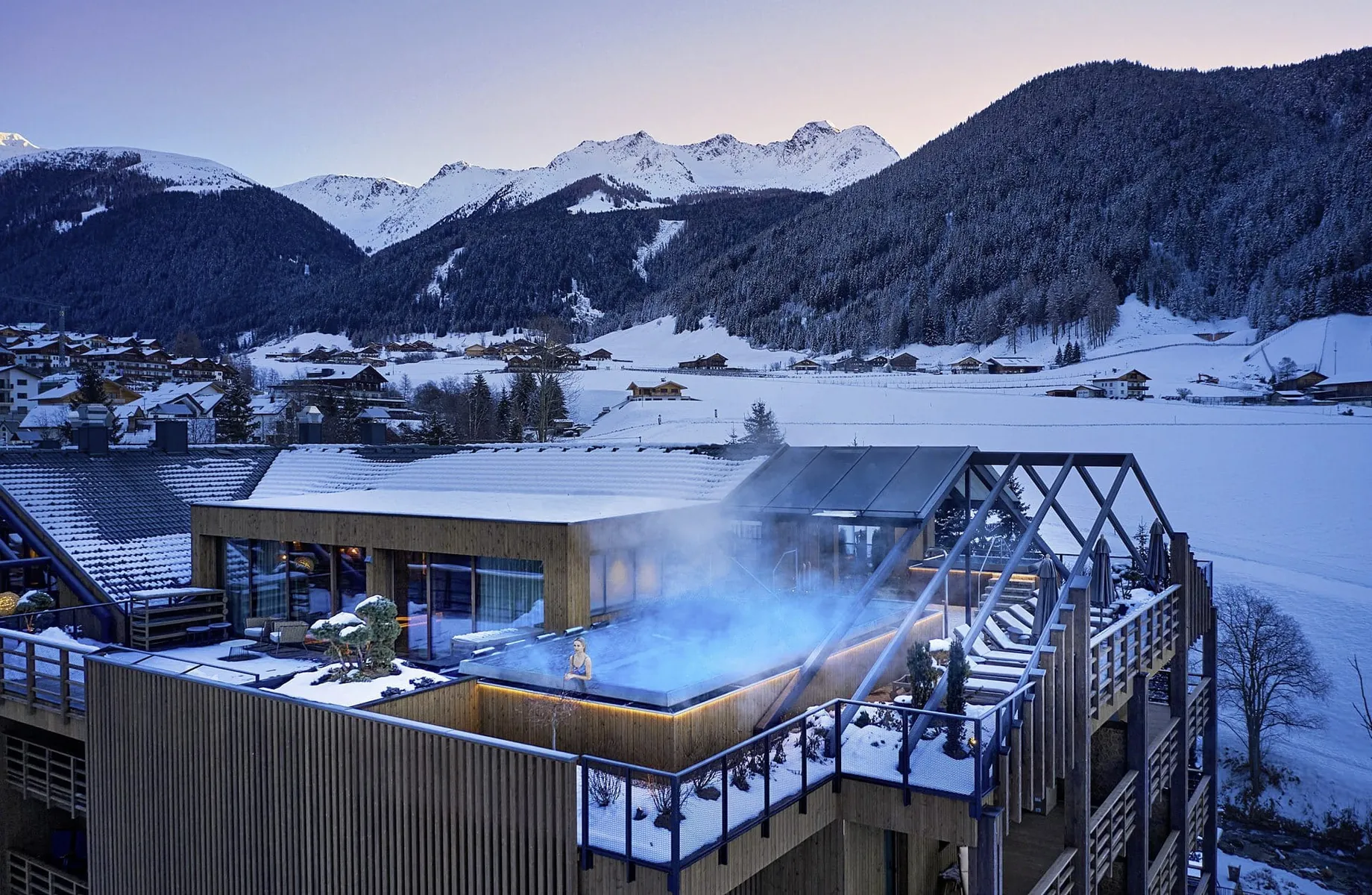 Außenansicht des Hotel Quelle Nature Spa Resort mit beheiztem Außenpool auf der Dachterrasse, umgeben von schneebedeckten Bergen und Winterlandschaft