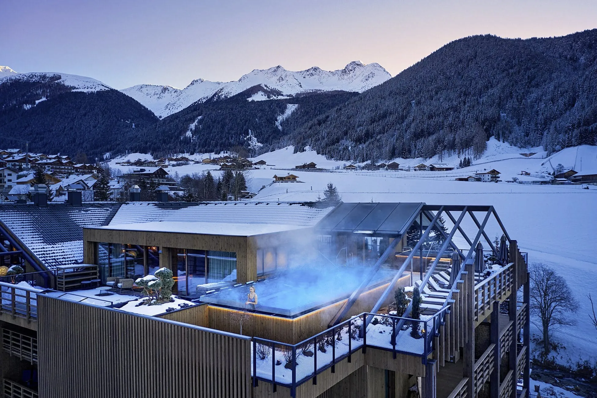 Außenansicht des Hotel Quelle Nature Spa Resort mit beheiztem Außenpool auf der Dachterrasse, umgeben von schneebedeckten Bergen und Winterlandschaft