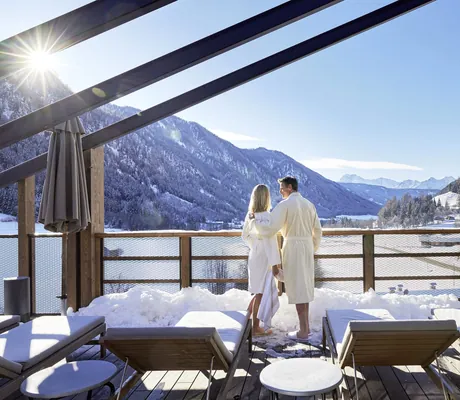 Paar in Bademänteln steht auf der Holzterrasse des Hotel Quelle Nature Spa Resort mit Liegen und Sonnenschirmen und blickt auf schneebedecktes Bergtal unter klarem Himmel