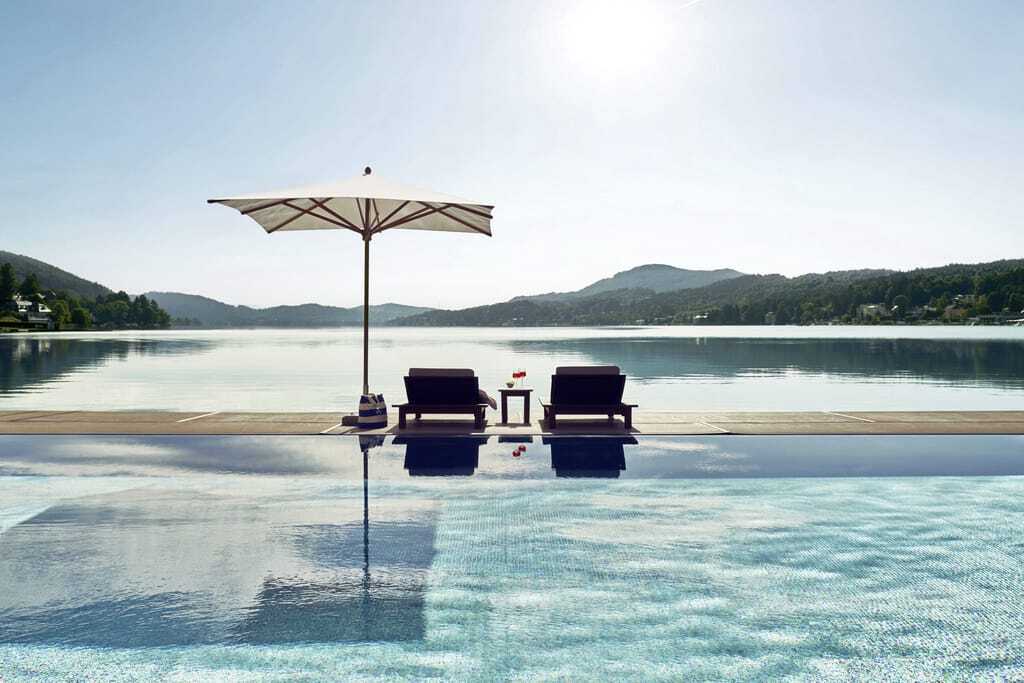 zwei Liegestühle unter einem Sonnenschirm am Outdoorpool im Falkensteiner Schlosshotel Velden mit Übergang zum See