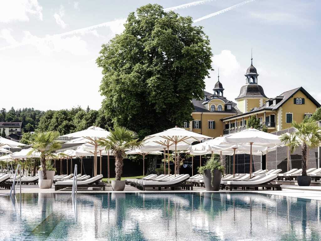 weiße Sonnenliegen unter weißen Sonnenschirmen am Outdoorpool des Falkensteiner Schlosshotel Velden mit Blick auf das Schloss