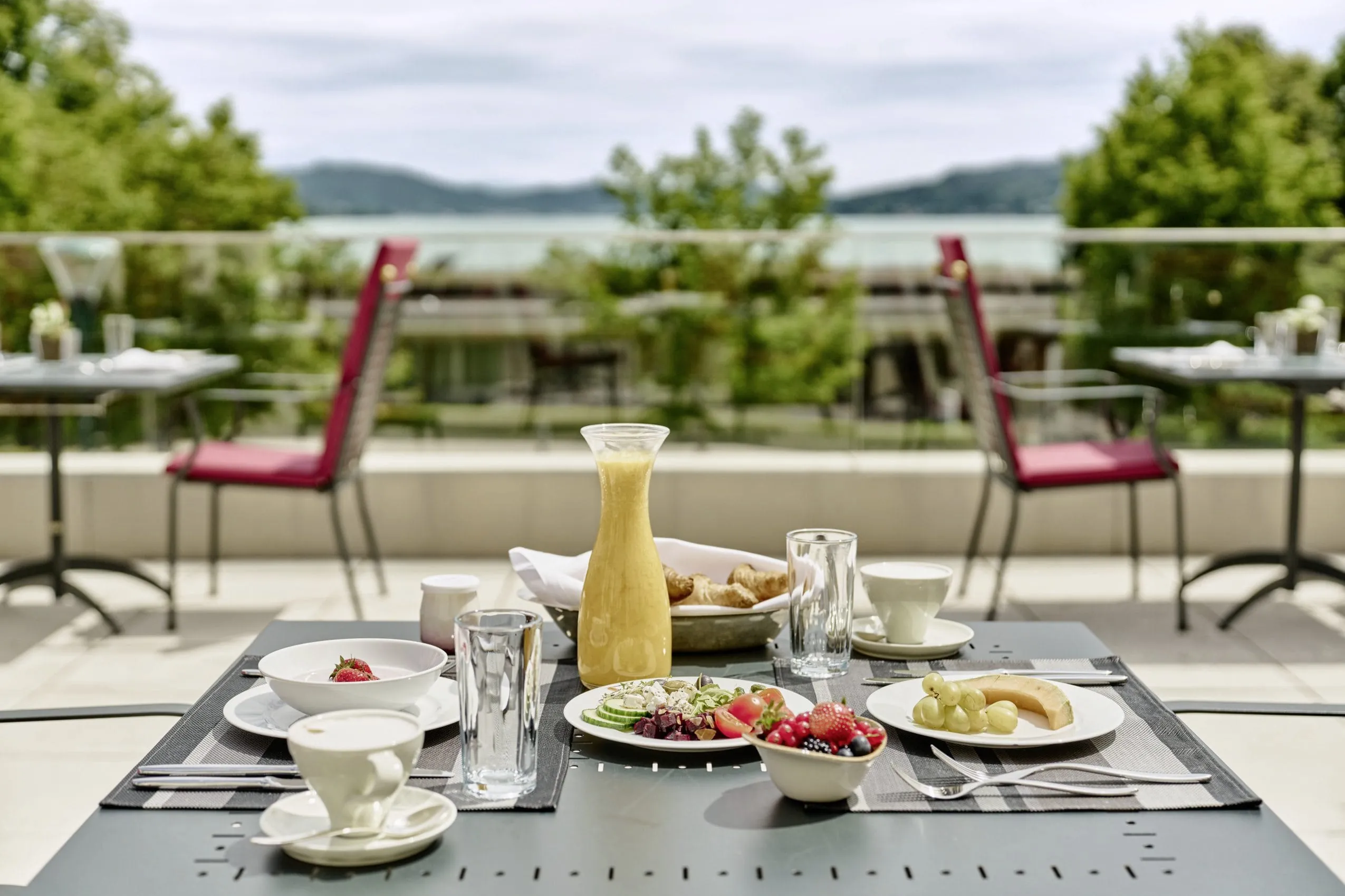 Fr&uuml;hst&uuml;ck auf der Terrasse des Falkensteiner Schlosshotel Velden mit rot gepolsterten St&uuml;hlen und Blick auf den W&ouml;rthersee