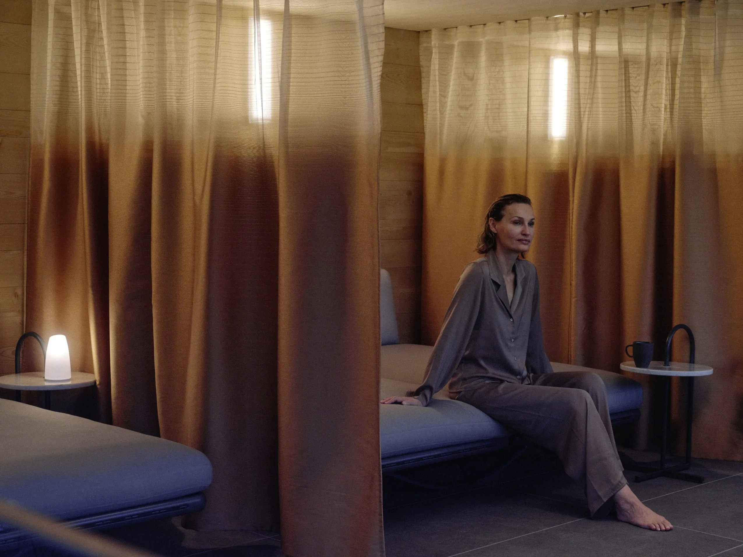 Frau in braunem Satinanzug sitzt auf Ruheliege zwischen Vorh&auml;ngen im Huus Quell