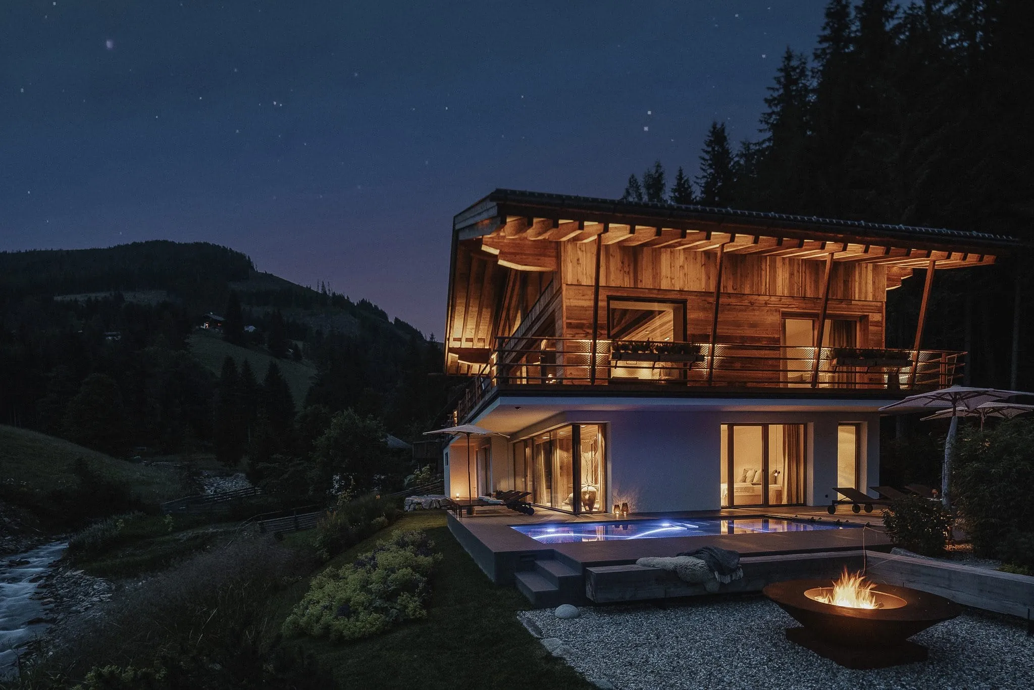 Modernes Chalet Falk aus Holz mit beleuchteter Terrasse und großen Glasfronten, umgeben von Wald und Hügeln, mit Pool und Feuerstelle im Vordergrund bei Nacht