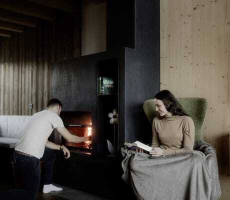 Mann schürt Ofen an in Kessler's Mountain Lodge während Frau mit Decke im Sessel ein Buch liest