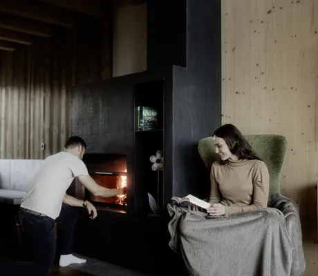 Mann schürt Ofen an in Kessler's Mountain Lodge während Frau mit Decke im Sessel ein Buch liest
