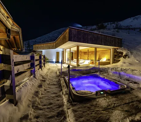 Beleuchteter Outdoor-Whirlpool des Chalet Stern in einer winterlichen Landschaft bei Nacht, im Hintergrund ein kleines Gebäude mit großen Glasfenstern und Ruheliegen im Innenbereich