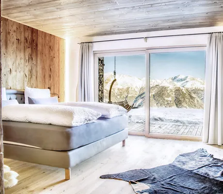 Modernes Schlafzimmer im Chalet Stern mit Doppelbett, Kamin, Fell auf dem Boden und bodentiefen Fenstern, die eine Aussicht auf verschneite Berge bieten