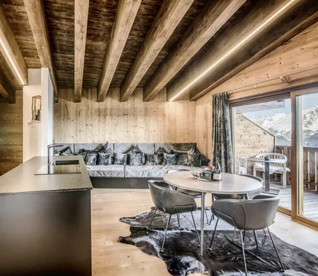 Gemütlicher Wohn- und Essbereich des Chalet Stern mit Holzbalkendecke, großem, grauen Sofa mit vielen Kissen, rundem Esstisch und Panoramafenster mit Bergblick