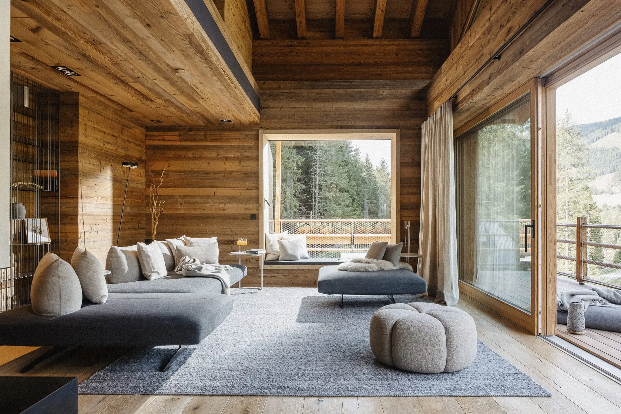 Moderner Wohnbereich mit Holzvert&auml;felung, gro&szlig;en Panoramafenstern, gem&uuml;tlichen grauen Sofas und Blick auf den Wald