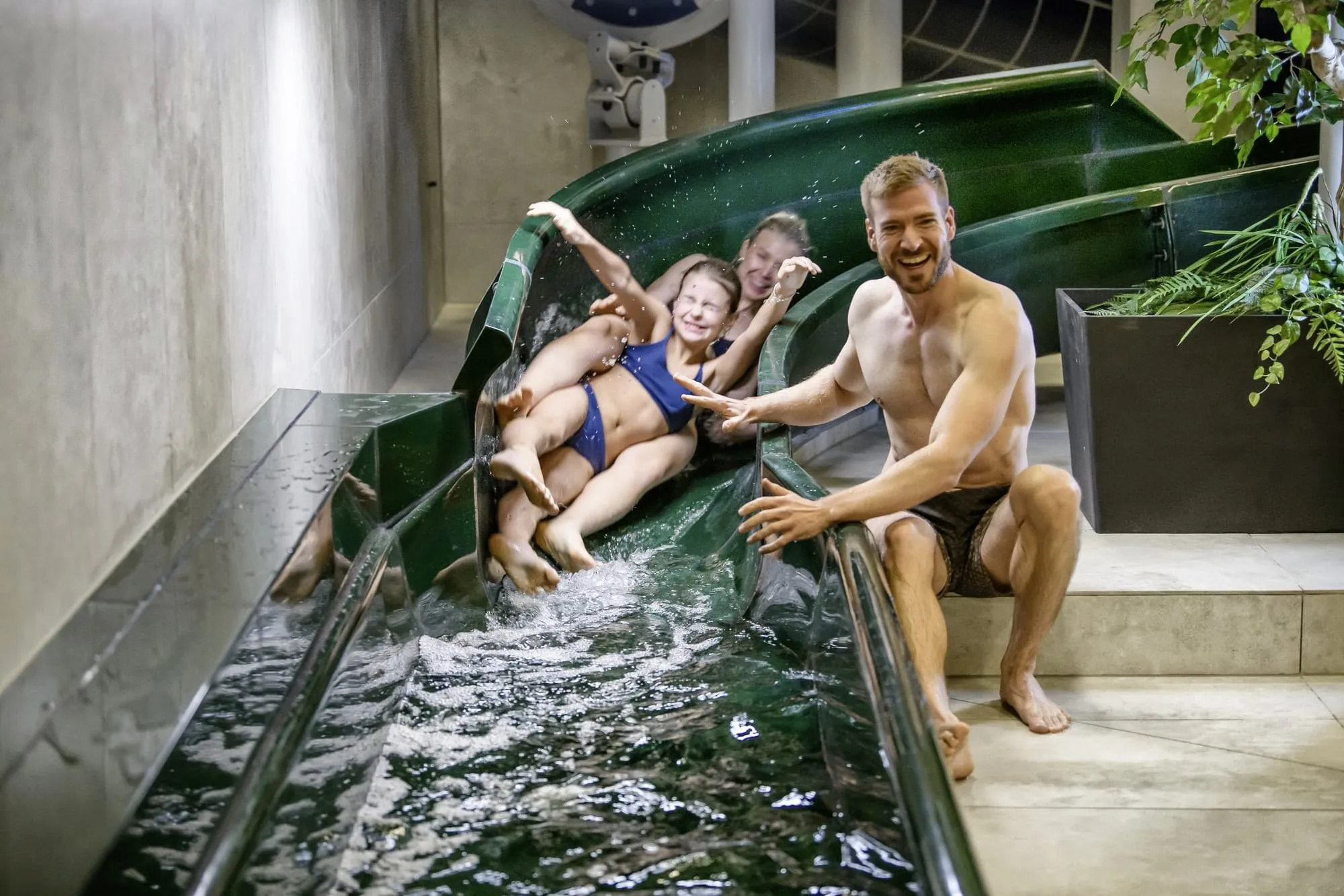 Mädchen und ihre Mutter rutschen lachend eine grüne Wasserrutsche im Familotel Zauchenseehof hinunter, der Vater sitzt daneben