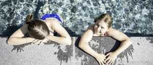 Zwei Mädchen lehnen am Rand eines Pools, umgeben von klarem Wasser im Das Mühlwald Quality Time Family Resort