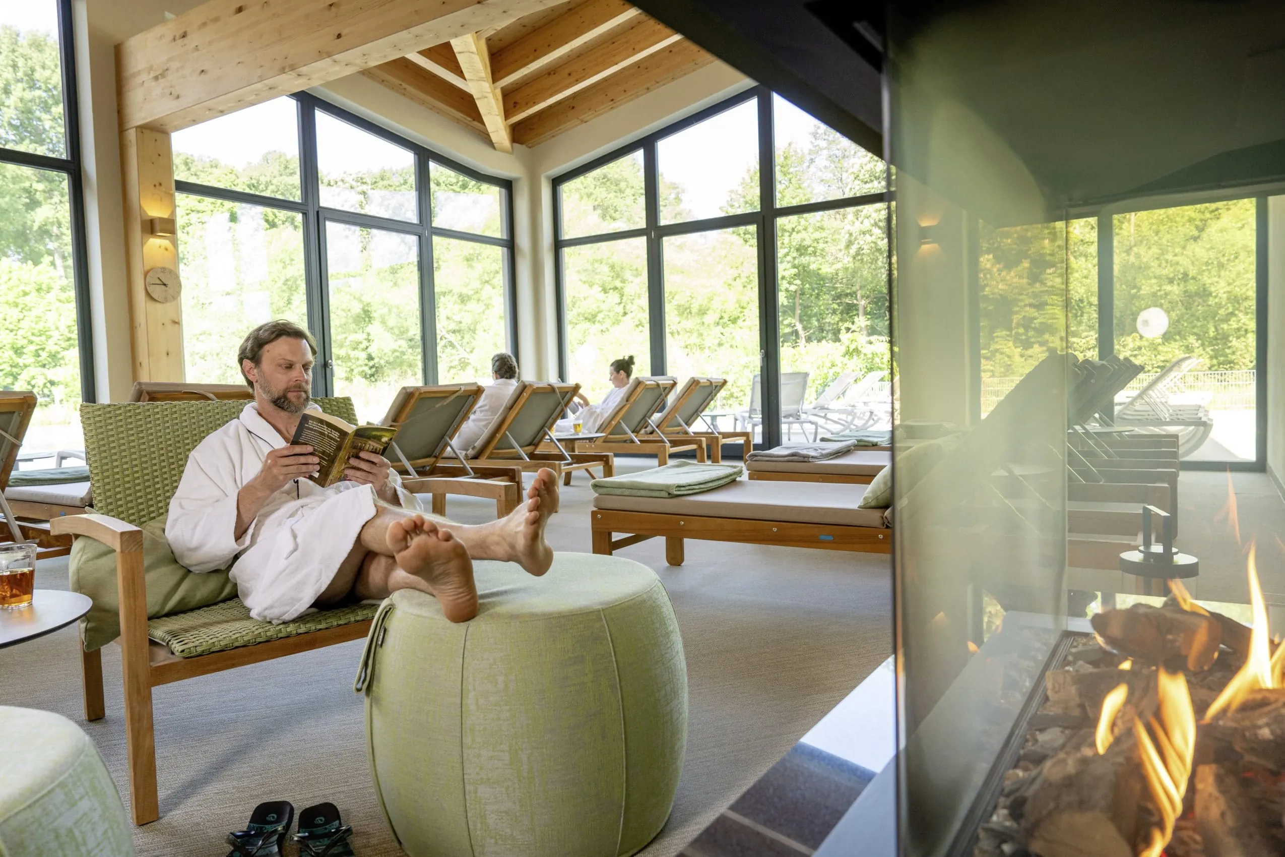 Mann in wei&szlig;em Bademantel sitzt mit hochgelegten F&uuml;&szlig;en mit Buch vor dem Kamin im Ruheraum des BEECH Resort Plauer See