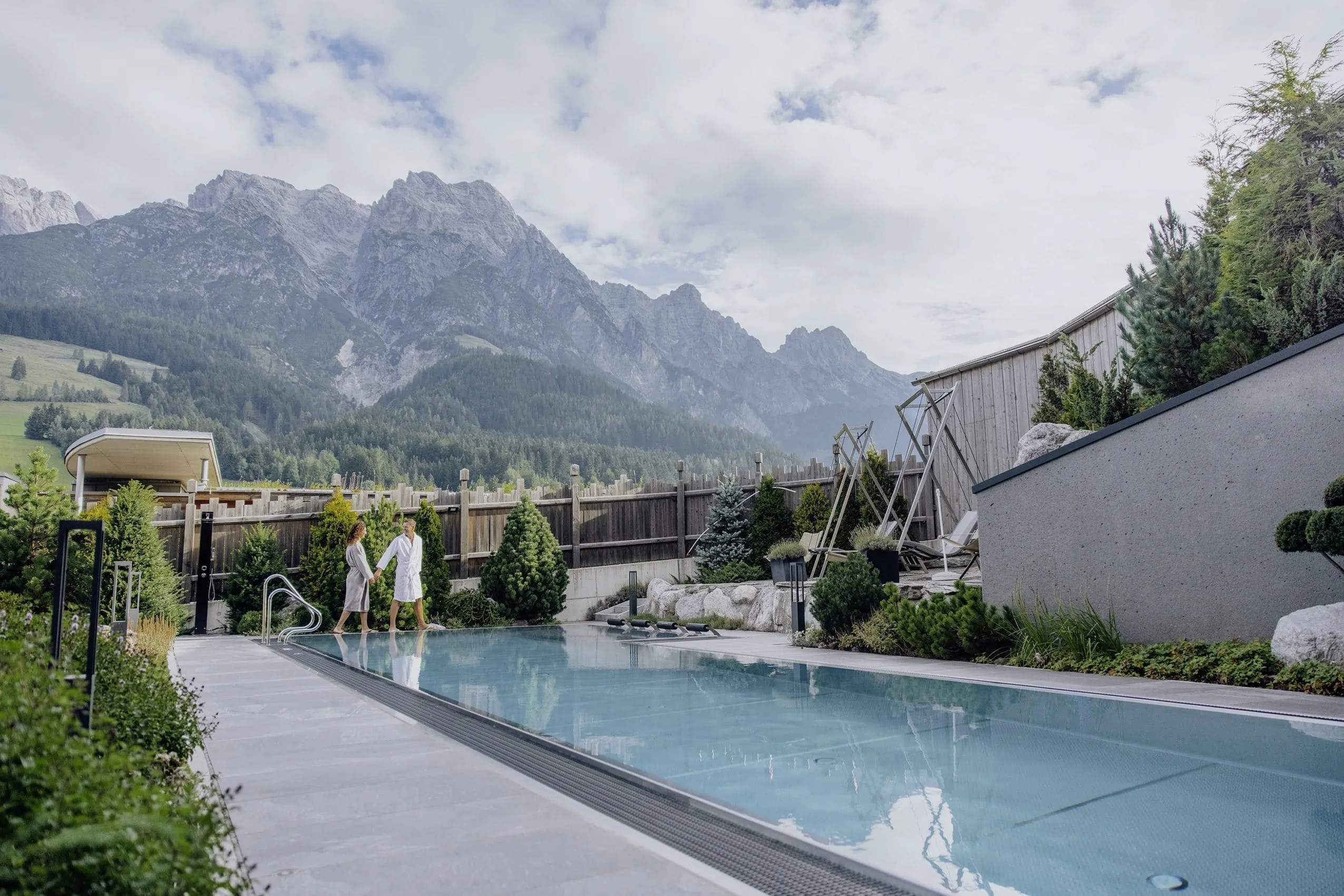 P&auml;rchen im Bademantel l&auml;uft um den Outdoor-Pool des Good Life Resort Riederalm mit Bergen im Hintergrund
