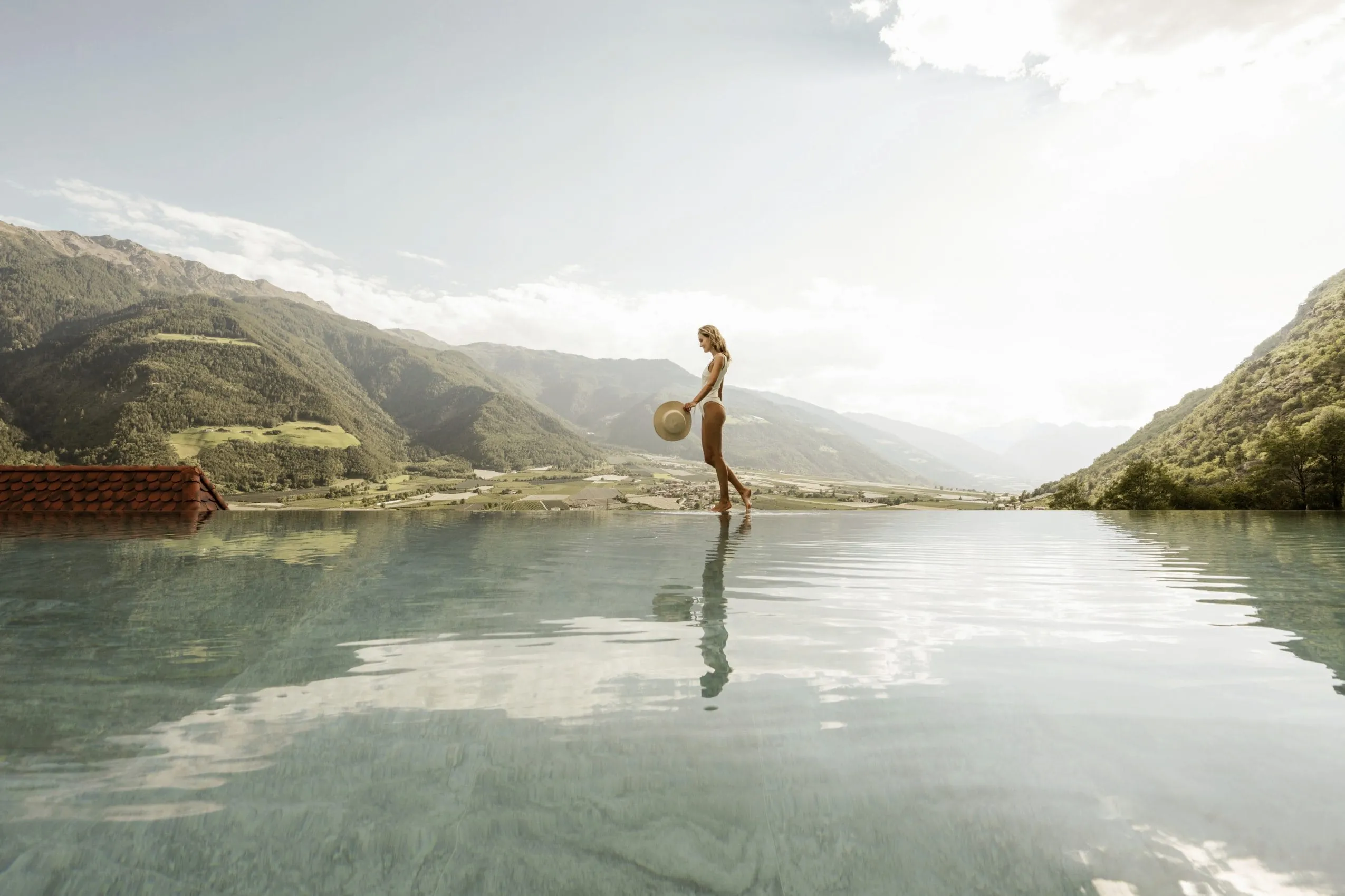 Frau in wei&szlig;em Badeanzug steht mit Hut in der Hand am Infinitypool des Preidlhof Luxury DolceVita Resort