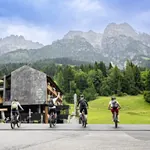 Außenansicht des mama thresl, Berge im Hintergrund, vier Fahrradfahrer im Vordergrund