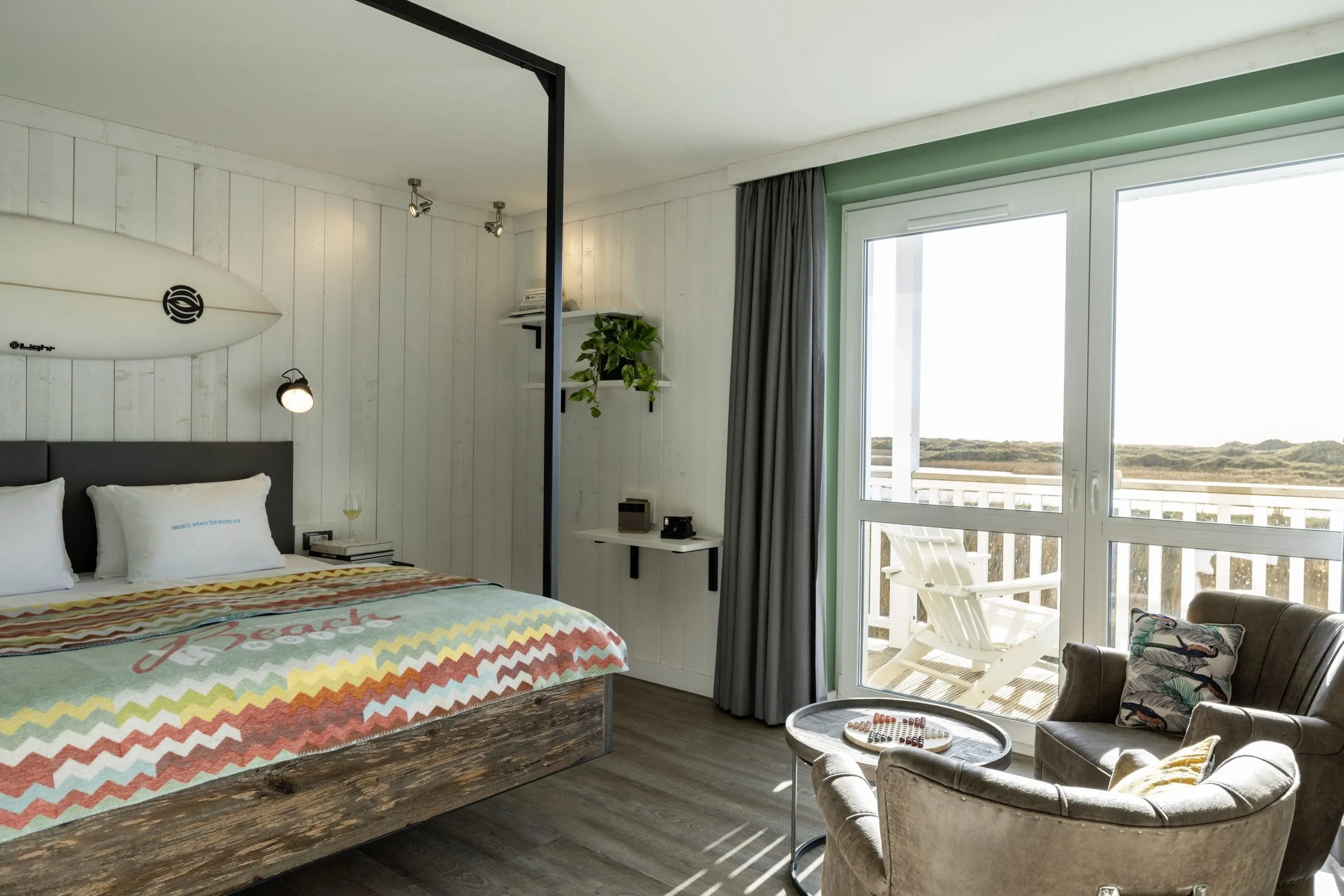 Blick in ein Zimmer des Beach Motel St. Peter-Ording mit Doppelbett, bunter Tagesdecke, Surfbrett als Wanddekoration, Sesseln und Balkon mit Ausblick