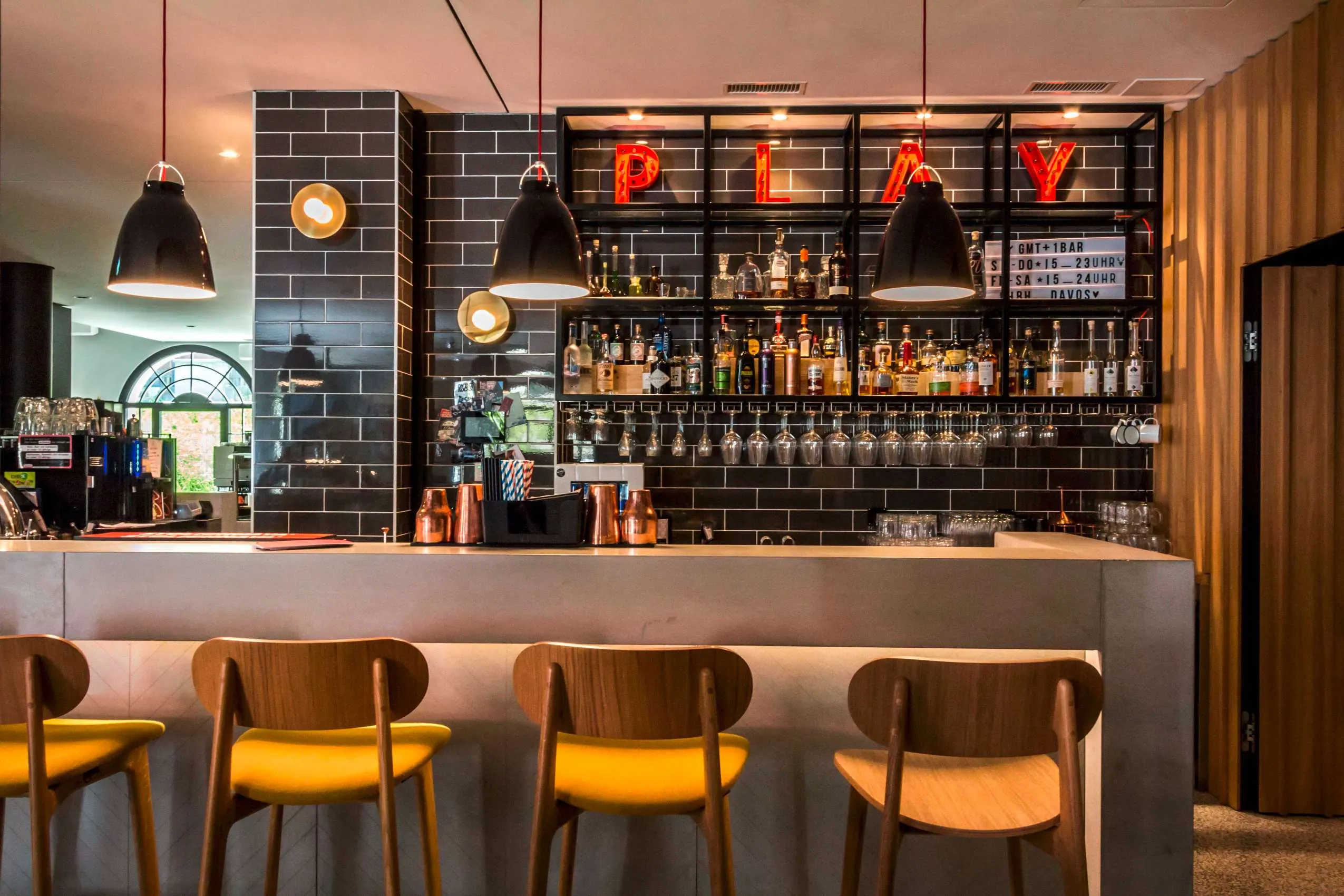 Moderne Bar im Hard Rock Hotel Davos mit schwarzer Wandfliesenoptik, beleuchteter "PLAY"-Dekoration, h&auml;ngenden Lampen, Holz-Barhockern und einer gut best&uuml;ckten Getr&auml;nkeauswahl