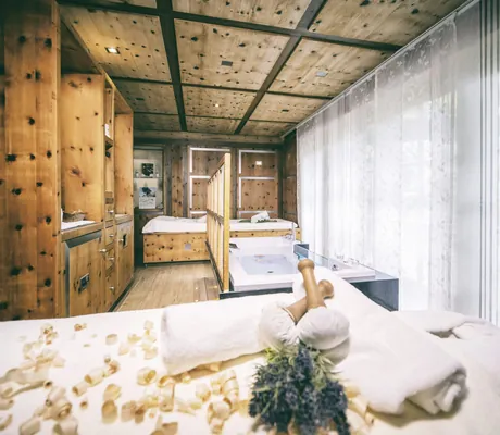 Behandlungsraum in der Wellnessresidenz Alpenrose mit Holzinterieur, Massagebett, Zirbenholzspänen und Blick auf helle Fenster mit Vorhängen