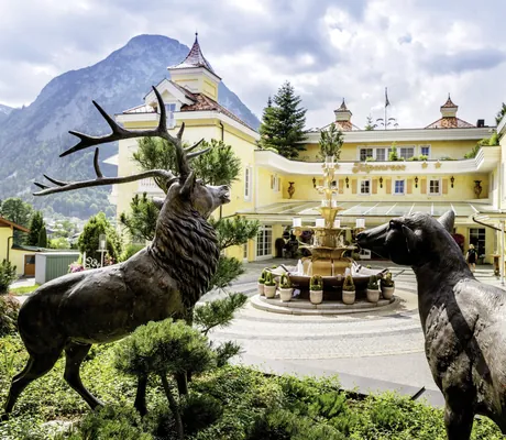 Außenansicht der Wellnessresidenz Alpenrose mit gelber Fassade, Brunnen und Bergkulisse im Hintergrund