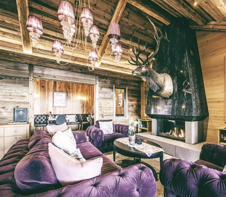 Stilvolle Lounge der Wellnessresidenz Alpenrose mit violetten Möbeln, Kamin und großem Hirschgeweih