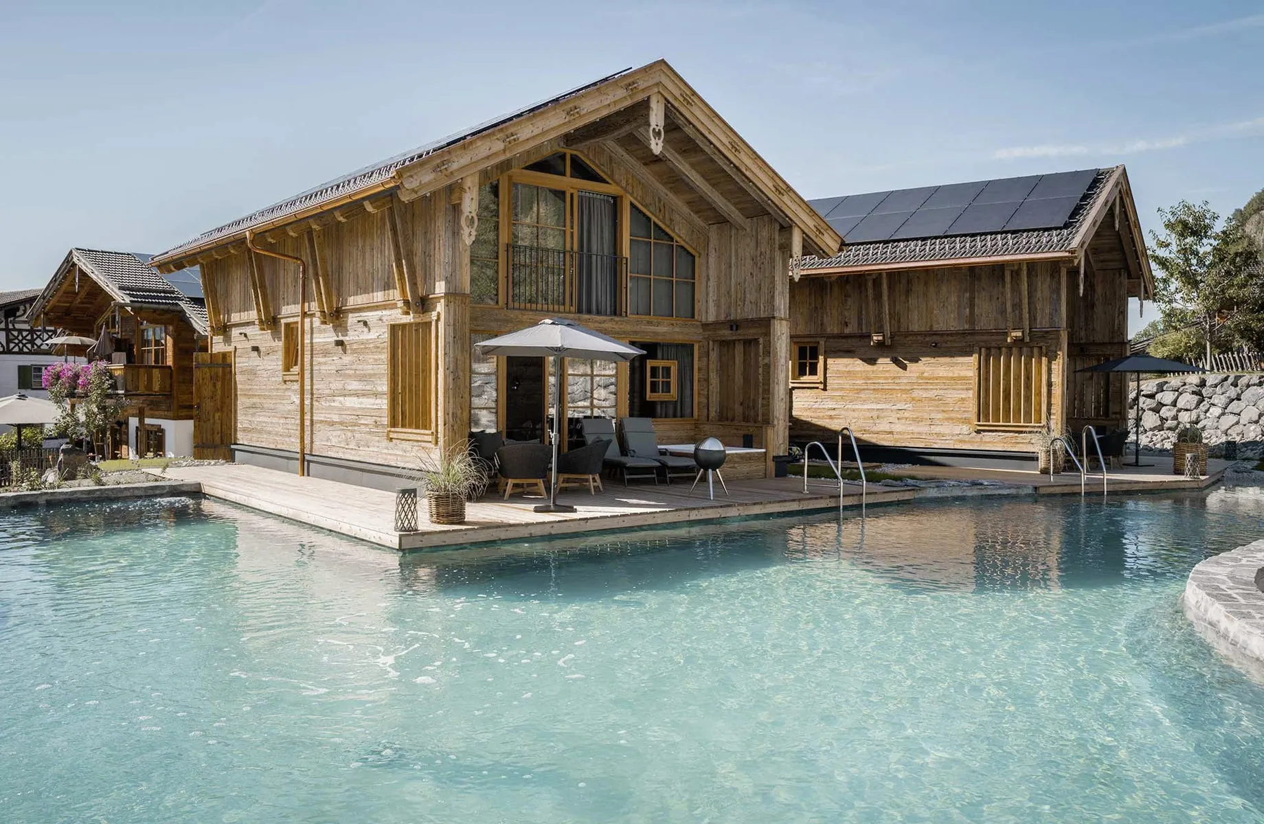 Sommerliche Außenansicht eines der Alpzitt-Chalets mit Holzfassade, Terrasse und Naturbadesee im Vordergrund