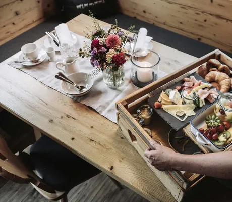 Frühstückskorb mit Aufschnitt, verschiedenem Obst, Backwaren, Blumen und gedecktem Tisch in den Alpzitt-Chalets