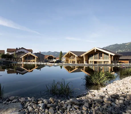 Gesamtansicht der Alpzitt-Chalets mit Natursee, mehreren Chalets und Berglandschaft im Hintergrund