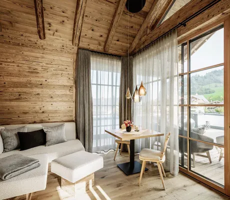 Gemütlicher Wohnbereich eines Alpzitt-Chalets mit Holzwänden, beigem Ecksofa, kleinem Esstisch und großen Fenstern mit Blick auf die Terrasse