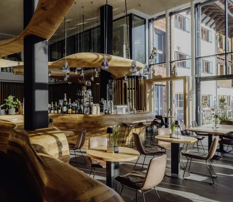Bar des Naturresort PURADIES mit geschwungenem Holzthekenbereich, mehreren runden Tischen und Stühlen, hängenden Lampen und großen Fenstern