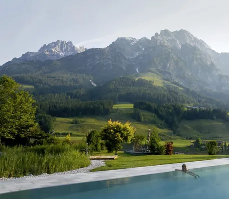Frau am Beckenrand des Outdoorpools im Naturresort PURADIES mit Sonnenliegen und Blick auf bewaldete Hügel und Bergpanorama