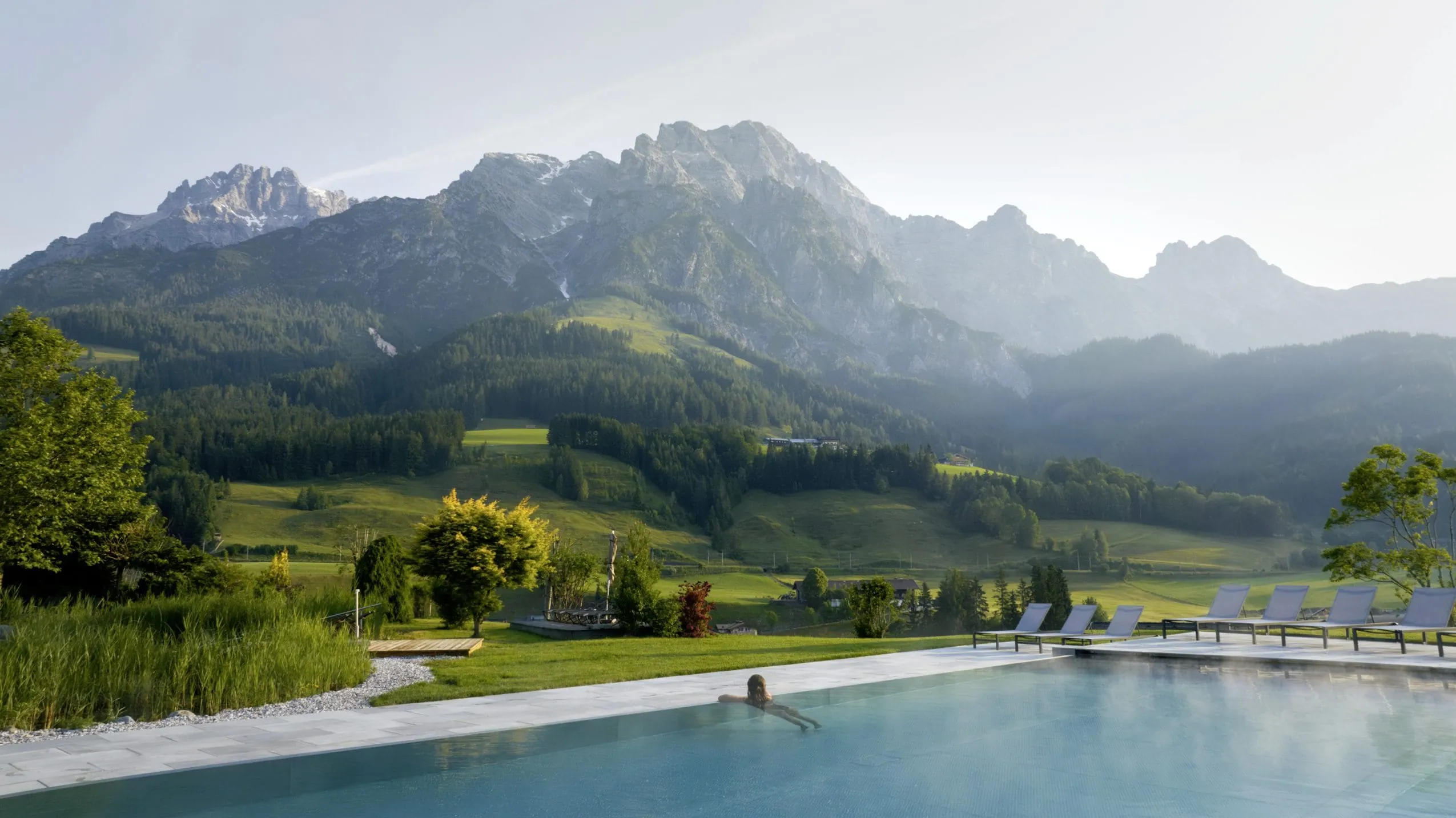 Frau am Beckenrand des Outdoorpools im Naturresort PURADIES mit Sonnenliegen und Blick auf bewaldete Hügel und Bergpanorama