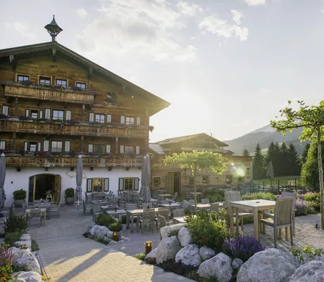 Sonnige Terrasse des Naturresorts PURADIES mit Holztischen, blühender Gartenlandschaft und Blick auf die Berge