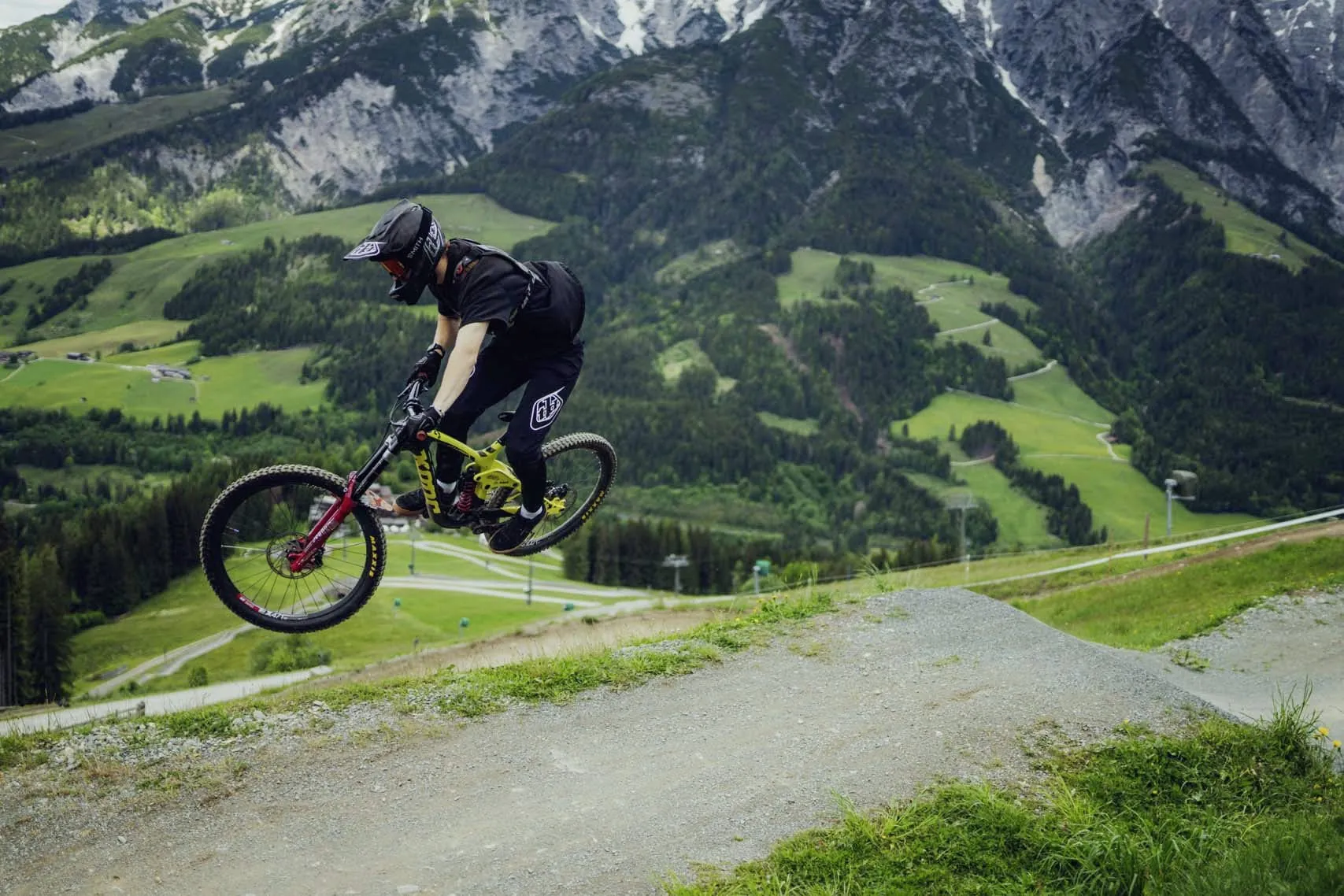 Mountainbiker in Aktion im Bikepark Leogang, umgeben von gr&uuml;nen Almwiesen und beeindruckenden Berggipfeln, im Hintergrund das Holzhotel Forsthofalm
