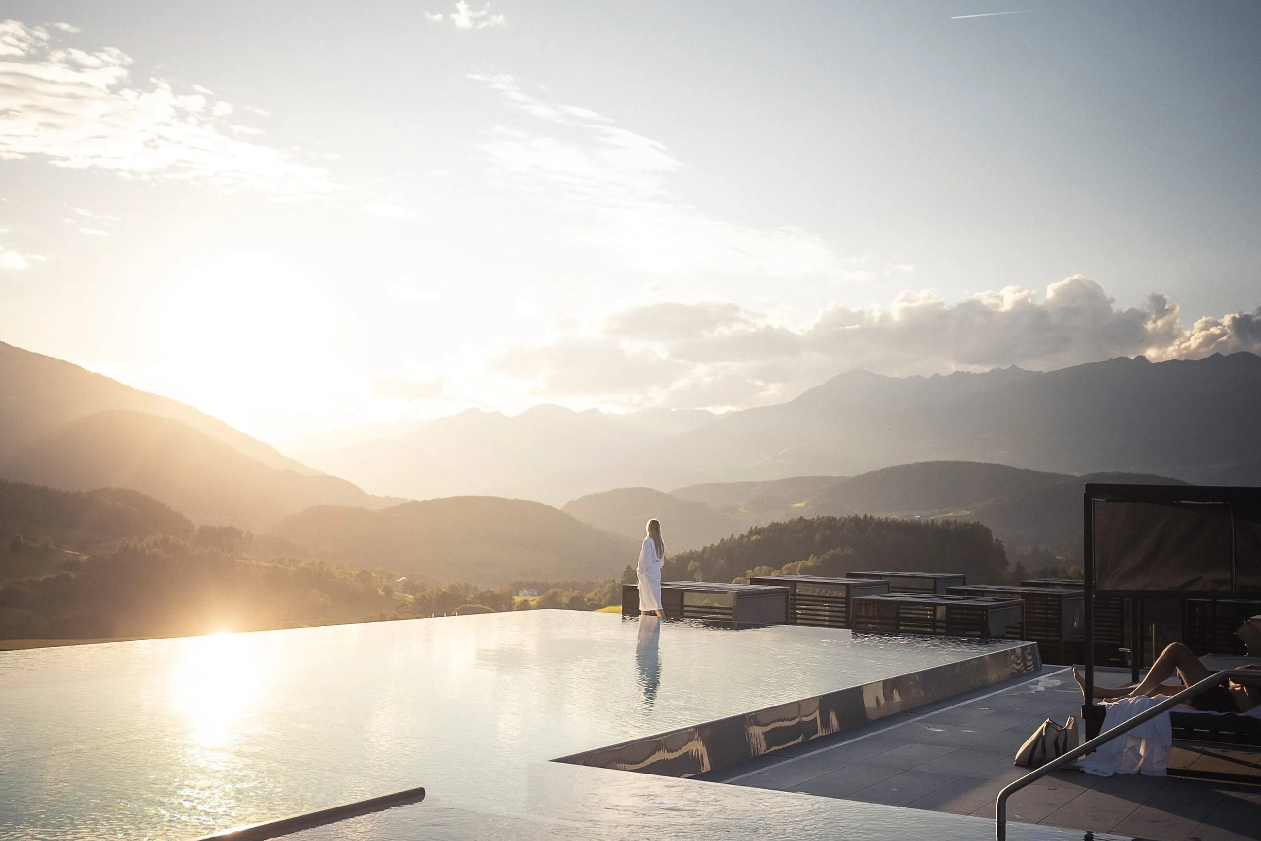 Frau im Bademantel l&auml;uft am Rand des Outdoor-Infinitypools im Hotel Winkler und blickt Richtung Berge