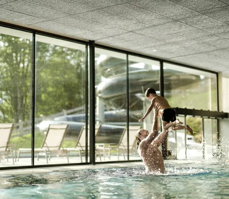 Vater spielt mit Kind im Indoorpool des Falkensteiner Family Hotels Montafon, große Fenster mit Blick nach draußen
