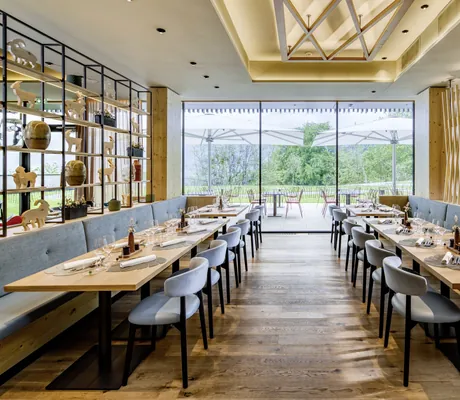 Modernes Restaurant im Falkensteiner Family Hotel Montafon mit Panoramafenstern, grauen Sitzmöbeln, Holztischen und -boden