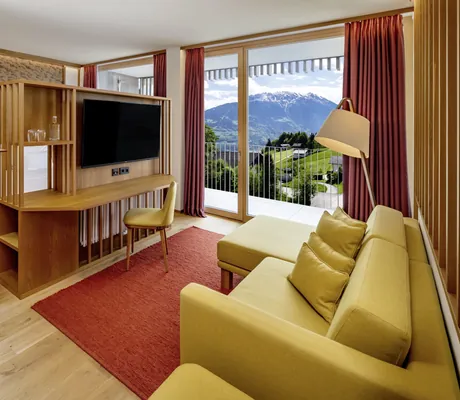 Zimmer im Falkensteiner Family Hotel Montafon mit Holzverkleidung, senfgelbem Sofa, rotem Teppich, Balkon und Bergblick