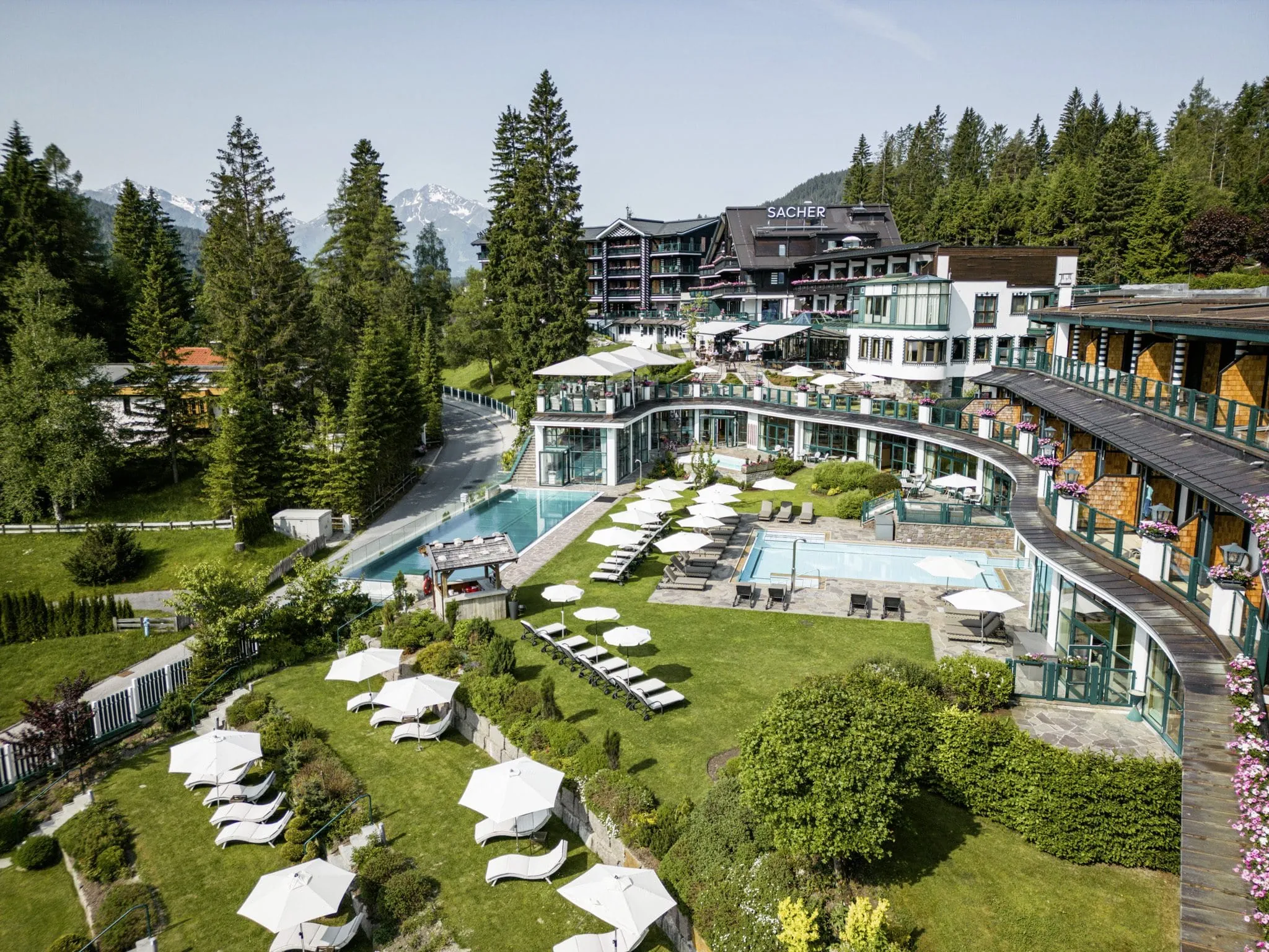 Blick auf das Gebäude des Alpin Resort Sacher Seefeld – Tirol im Sommer mit zwei Outdoorpools und großer Liegewiese