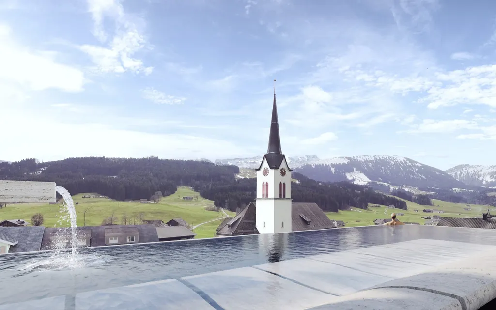 Weitblick auf Kirche und schneebedeckte Berge vom Sky-Infinitypool des HUUS Quell.