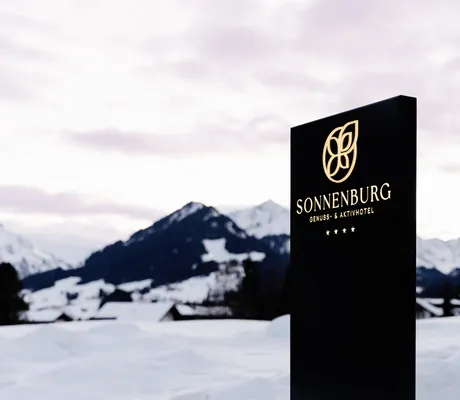 Schild mit Hotellogo vor einer Bergkulisse mit Schnee.