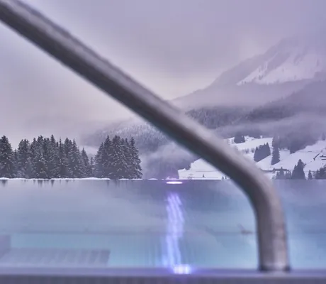 Blick auf den Infinitypool des Hotel Sonnenburg vor schneebedecktem Berg und Wäldern.