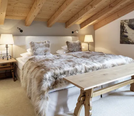 Gemütliches Schlafzimmer in einem der Alpenchalets des Sonnenalp Resorts mit Holzbalkendecke, Doppelbett mit Fell-Überwurf und Nachttischen