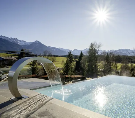 Outdoorpool einer NaturLodge des Sonnenalp Resorts mit Wasserfontäne, strahlender Sonne und Panoramablick auf grüne Landschaft und Berge