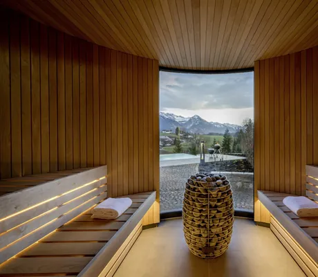Moderne Holzsauna einer NaturLodge des Sonnenalp Resorts mit Panoramafenster und Steinofen in der Mitte, Blick auf Berge und Außenbereich mit Pool
