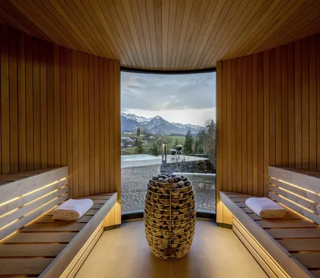 Moderne Holzsauna einer NaturLodge des Sonnenalp Resorts mit Panoramafenster und Steinofen in der Mitte, Blick auf Berge und Außenbereich mit Pool