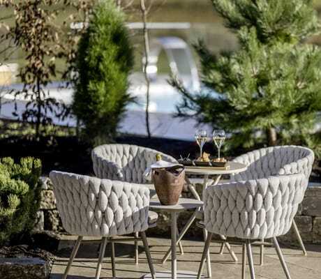 Terrasse einer NaturLodge des Sonnenalp Resorts mit vier gepolsterten Stühlen, gedecktem Tisch, Champagner und Snacks, umgeben von Pflanzen und Pool im Hintergrund